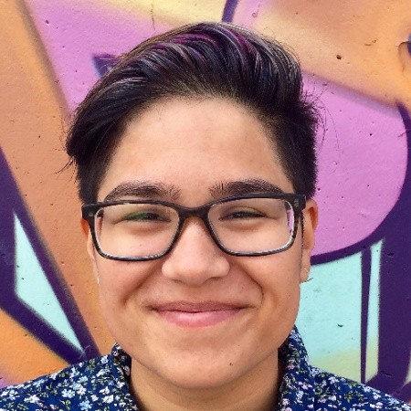 a headshot of Zo's face while they stand in front of a graffiti wall. Their combed over hair has streaks of dark purple He wears thick rectangle framed glasses and has a slight smile at the camera with a blue floral collar shirt.