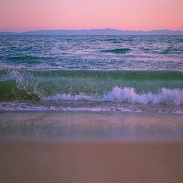 View of ocean waves crashing into shore during a pink hue sunset. The ocean waves are a greenish pale hue.