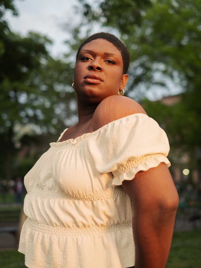 A non-binary trans woman posing in a park on a sunny day while wearing a cream shoulder shirt with ruffle ends.