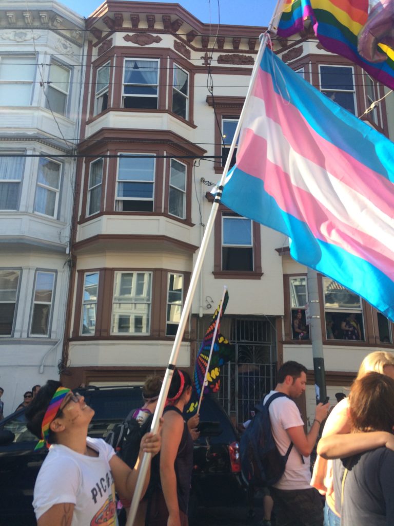 A nonbinary person with a rainbow bandana on their head holding a telescope pole with the trans flag and rainbow flag. They are marching down the street during SF Pride.